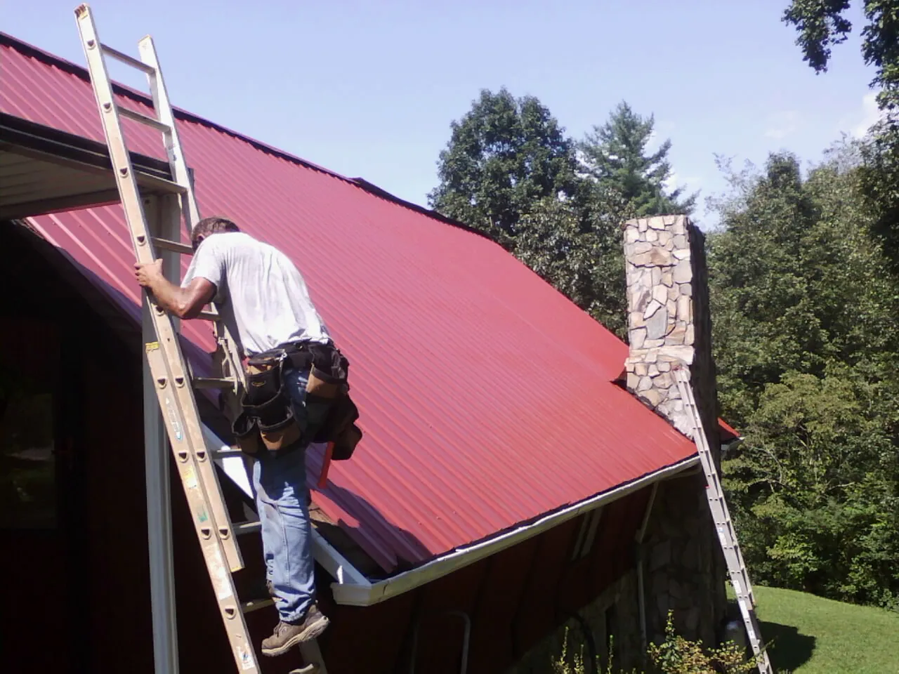 Roofer on a ladder beside a red metal roof and stone chimney — Soffit & Fascia Repair by Roofing Brothers in Lowellville